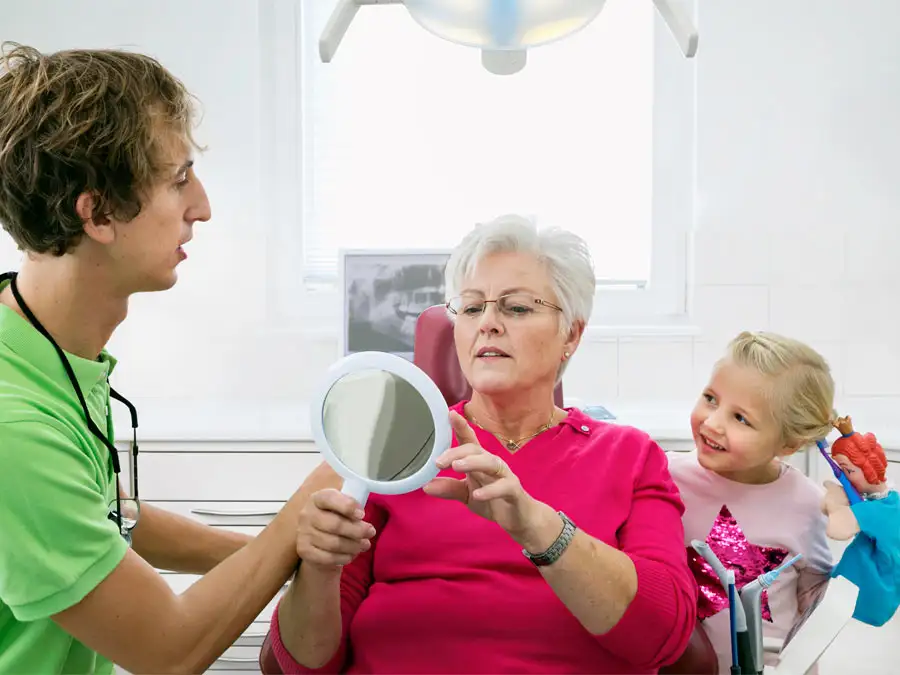 Dr. med. dent. Johannes Haderthauer zeigt Patientin im Spiegel das Behandlungsergebnis und ein Kind mit Prinzessinnenpuppe schaut zu. Praxis an der Elz – Zahnheilkunde & Kieferorthopädie, Teningen