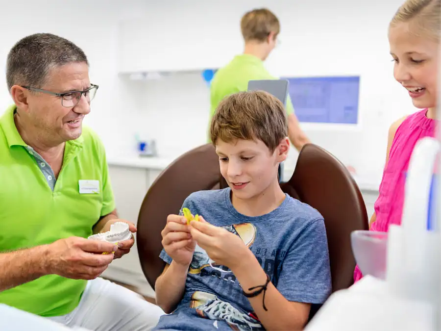 Kieferorthopäde Dr. Meschenmoser erklärt die Funktionsweise einer losen Spange einem Jungen und einem zuschauendem Mädchen. Er hält ein Gipsmodell in der Hand.